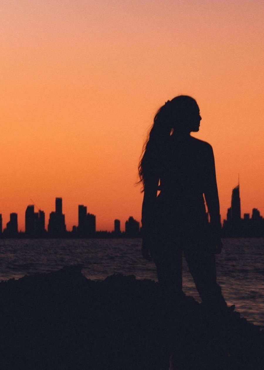 silhouette of woman looking right against golden hour sunset drop behind city skyline across lake